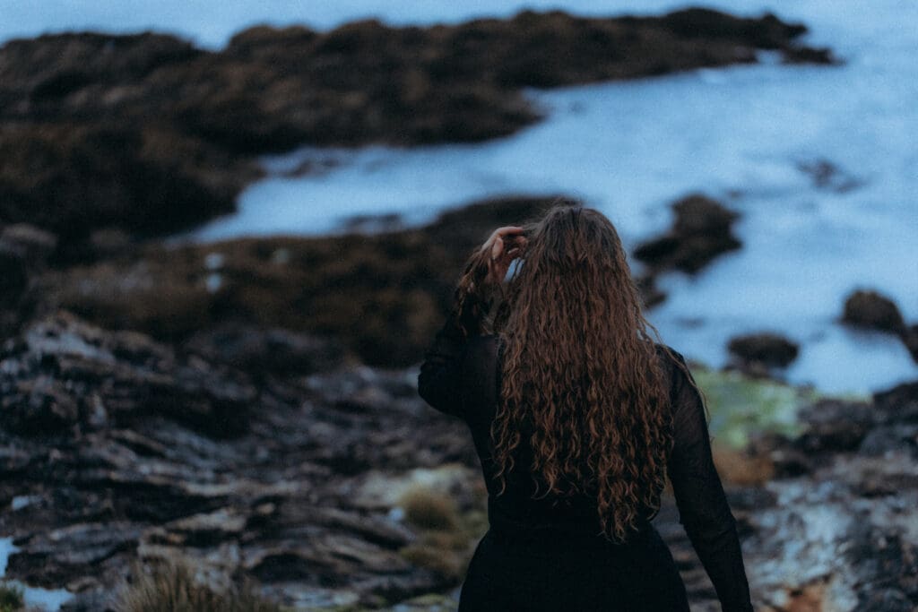 Femme de dos marchant sur une plage de roches, cheveux au vent, photographie cinématographique
