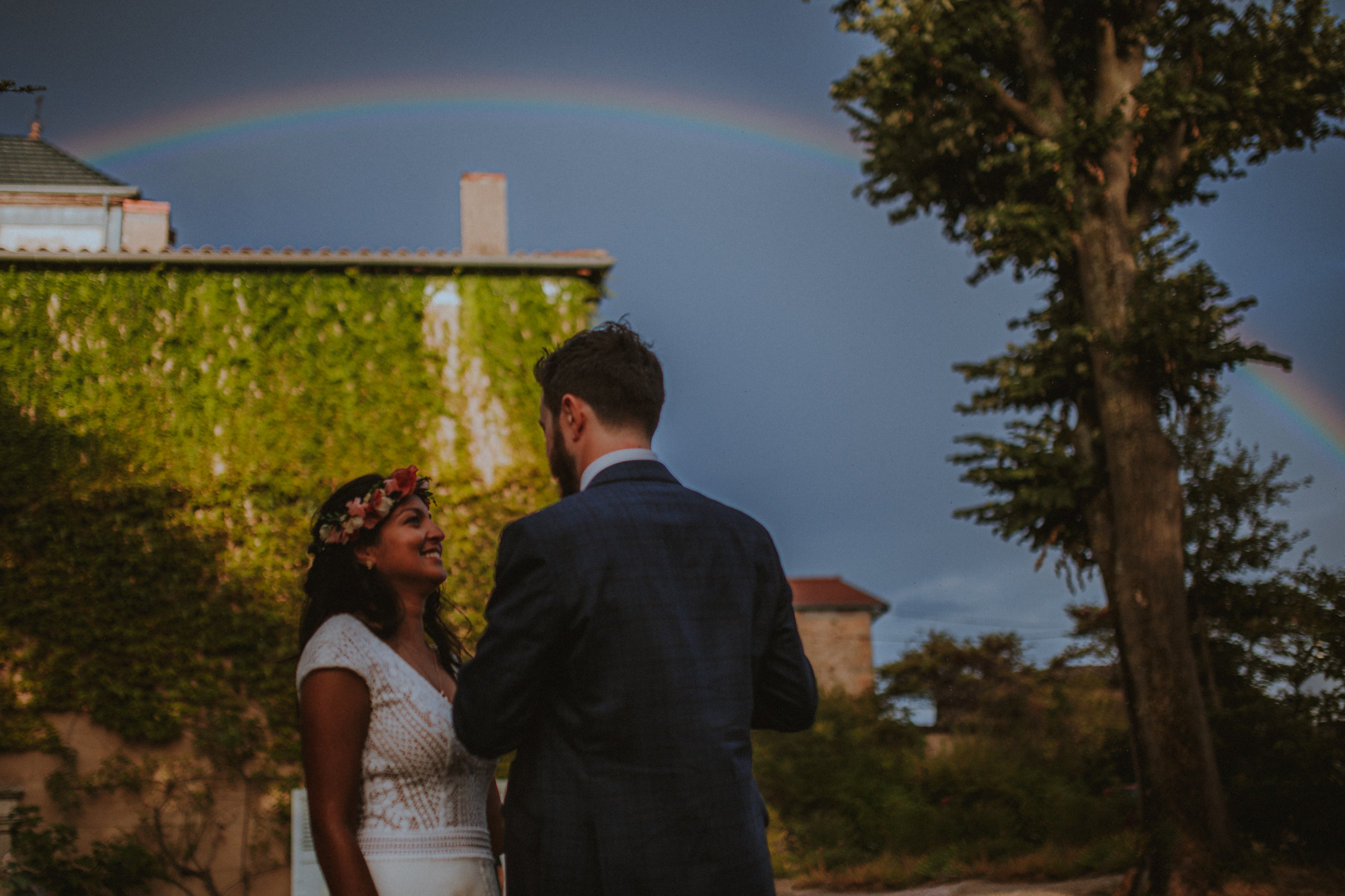 Mariés qui marchent dans un jardin avec un arc-en-ciel dans le ciel
