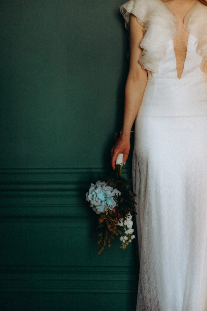Mariée de dos devant un mur vert, bouquet à la main, avant la cérémonie