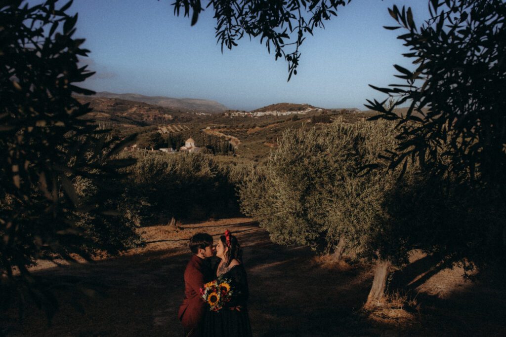 Photographe mariage Crète, Elodie & Marvin enlacés dans un champ d’oliviers, village et chapelle au loin