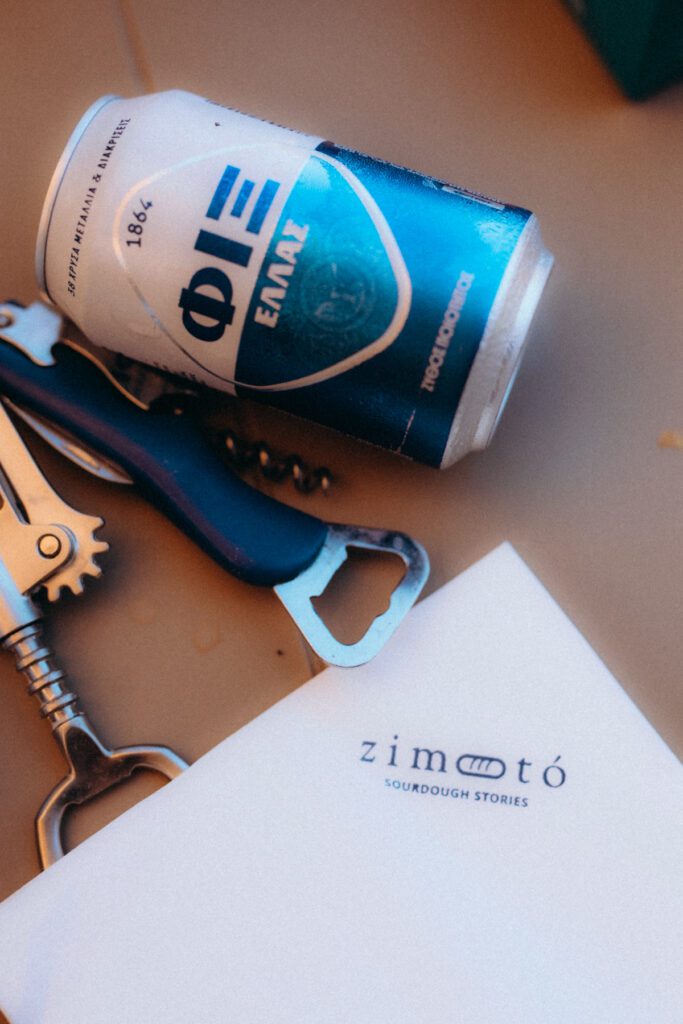 Canette de bière grecque, décapsuleur et papier à l’enseigne Zimotó posés sur une table